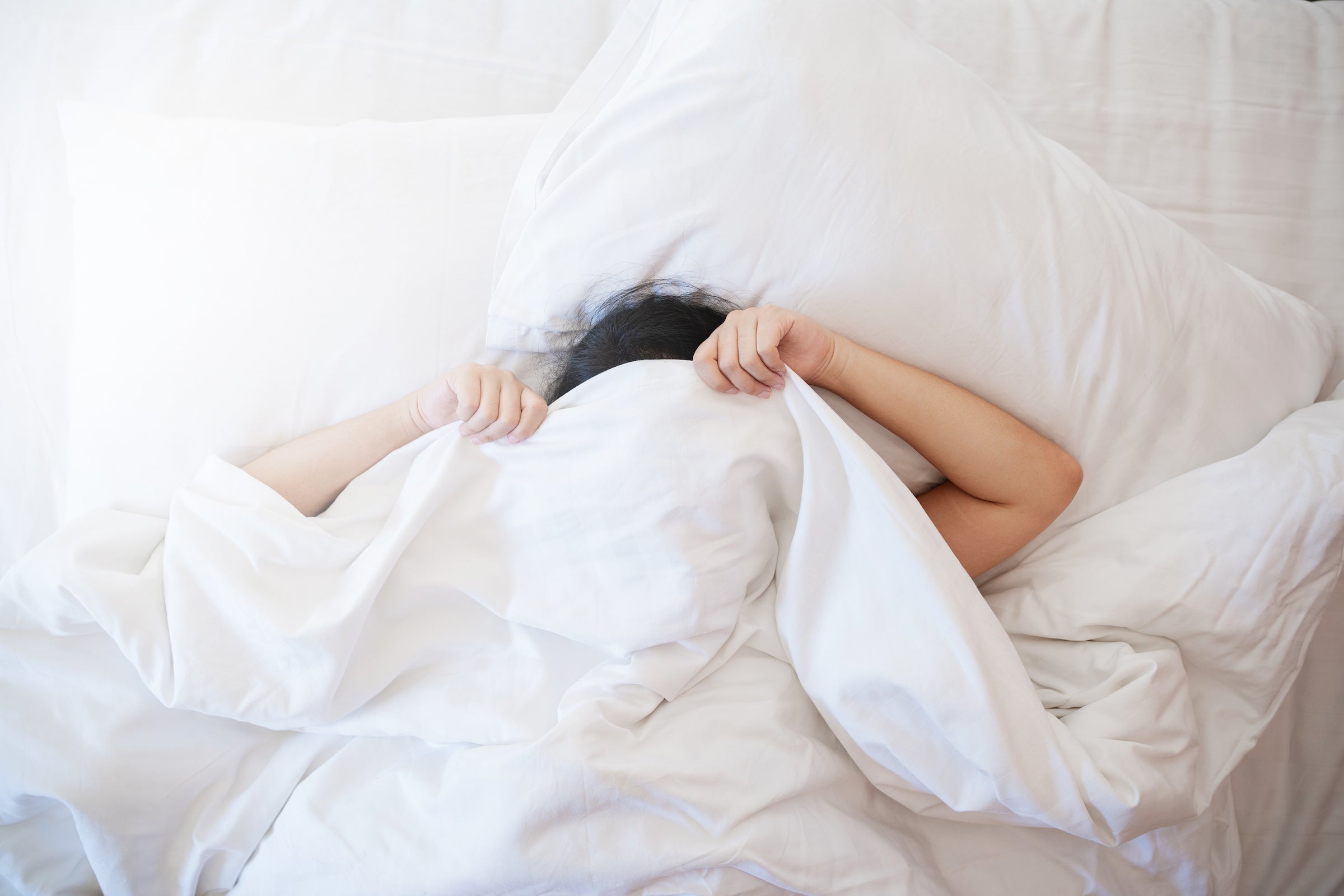 Ein dunkelhaarige Frau liegt im Bett und zieht sich die Bettdecke über den Kopf