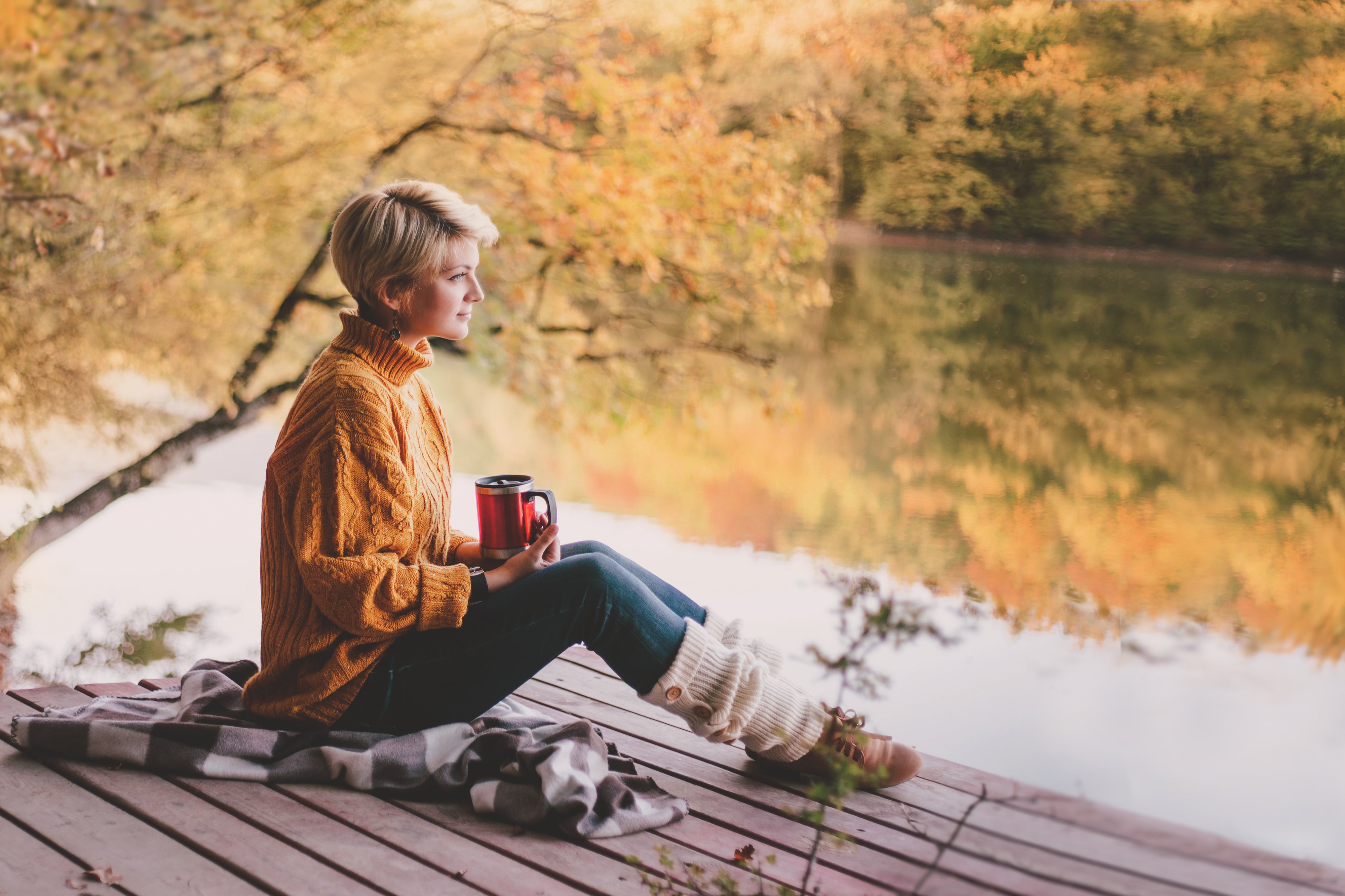 Eine blonde, kurzhaarige Frau sitzt an einem herbstlichen See und hält eine Thermoskanne