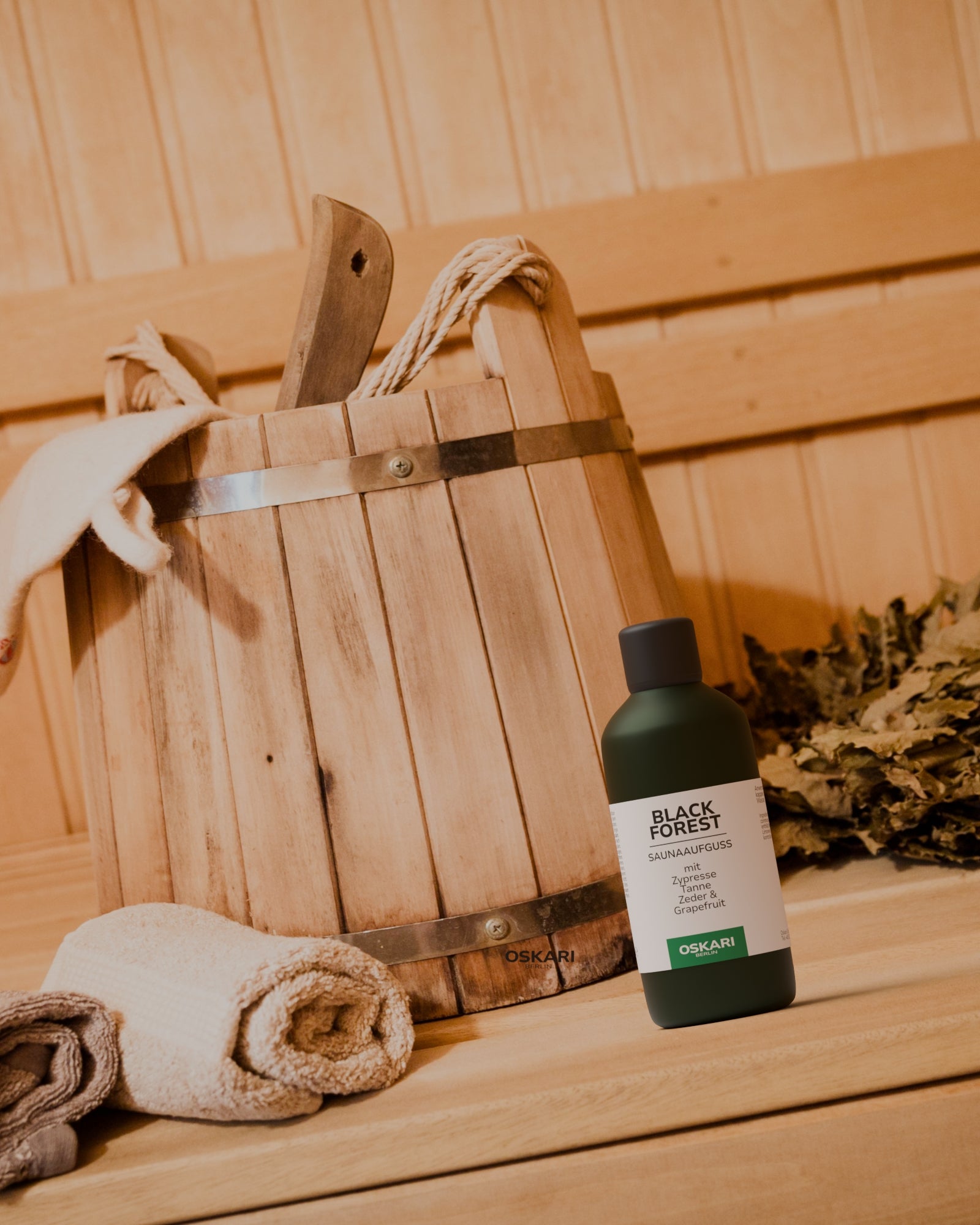 Grüne Flasche mit Saunaaufguss Black Forest in hölzerner Sauna vor Saunaaeimer aus Holz und Handtuch