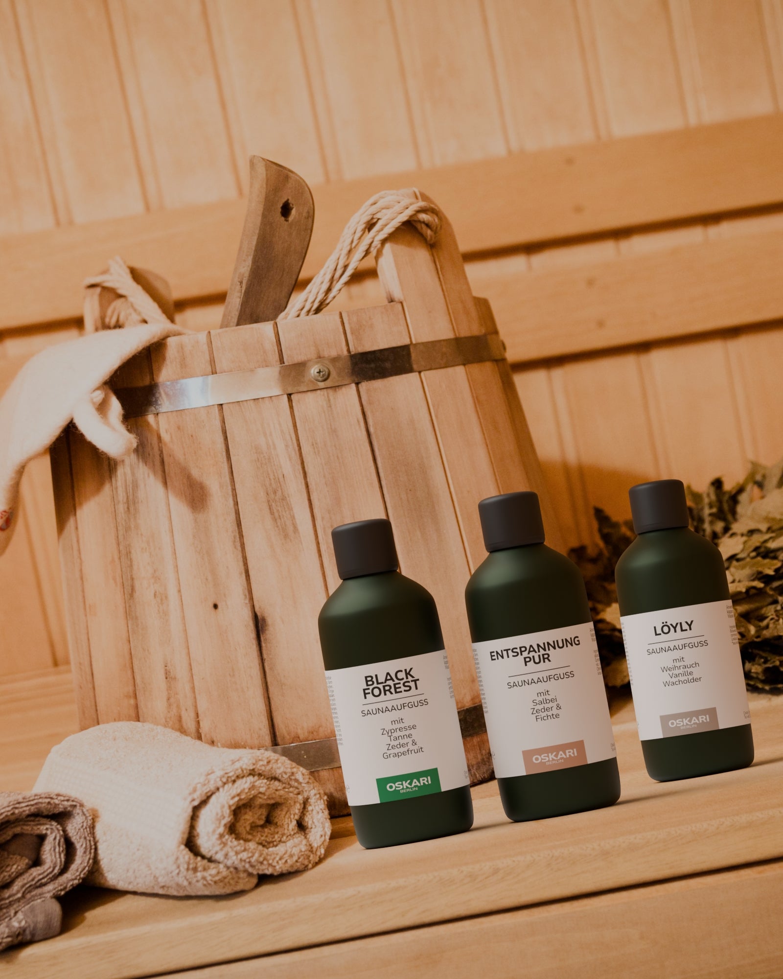 Die Saunaaufgüsse Black Forest, Entspannung Pur und Löyly stehend in einer Sauna vor Holzeimer mit Holzkelle und Handtuch