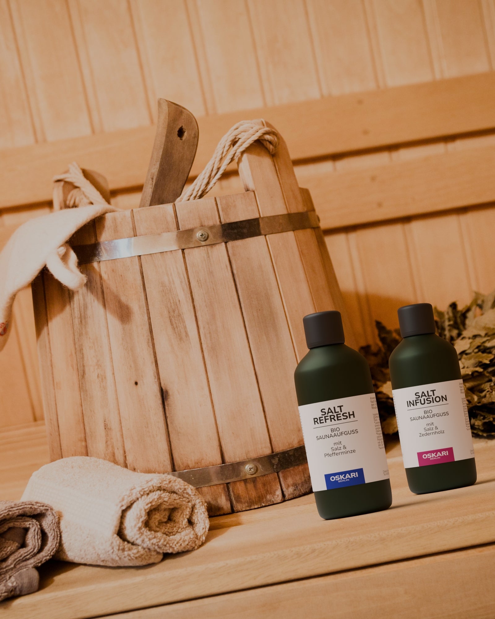 Die beiden Bio Salz-Saunaaufgüsse Salt Refresh und Salt Infusion in einer Sauna vor hölzernem Saunaeimer mit Handtuchrolle im Vordergrund