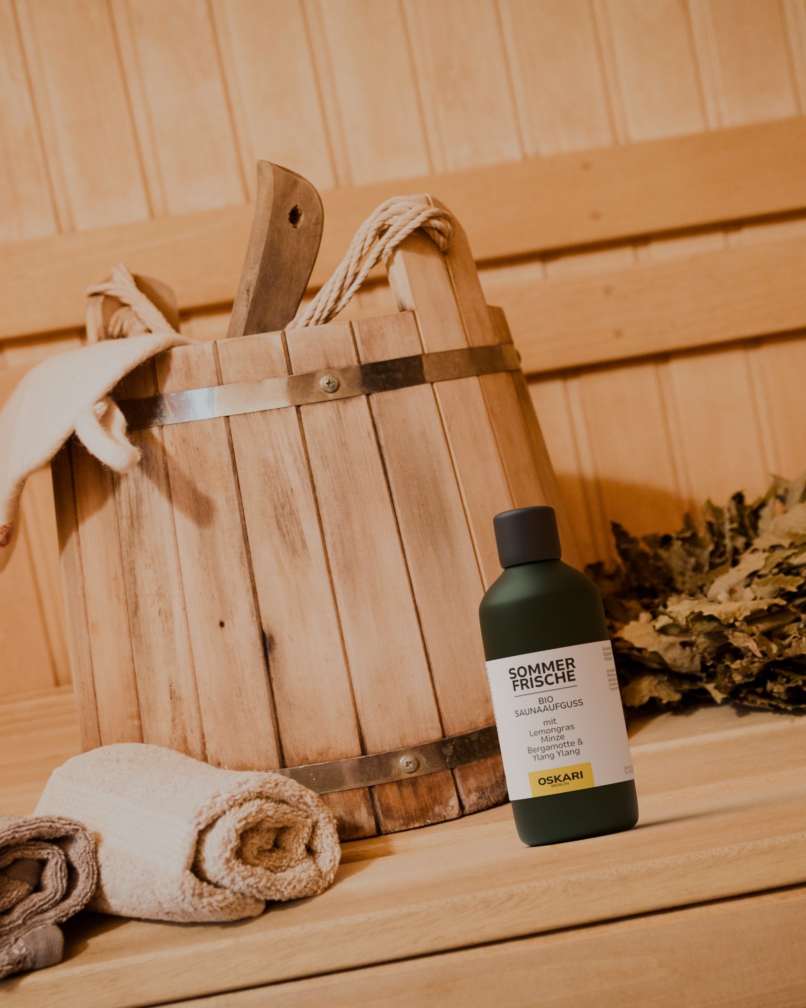 Der Oskari Bio Saunaaufguss Sommerfrische in der Grünglasflasche steht in einer Sauna mit einem Saunaeimer und Handtuch im Hintergrund