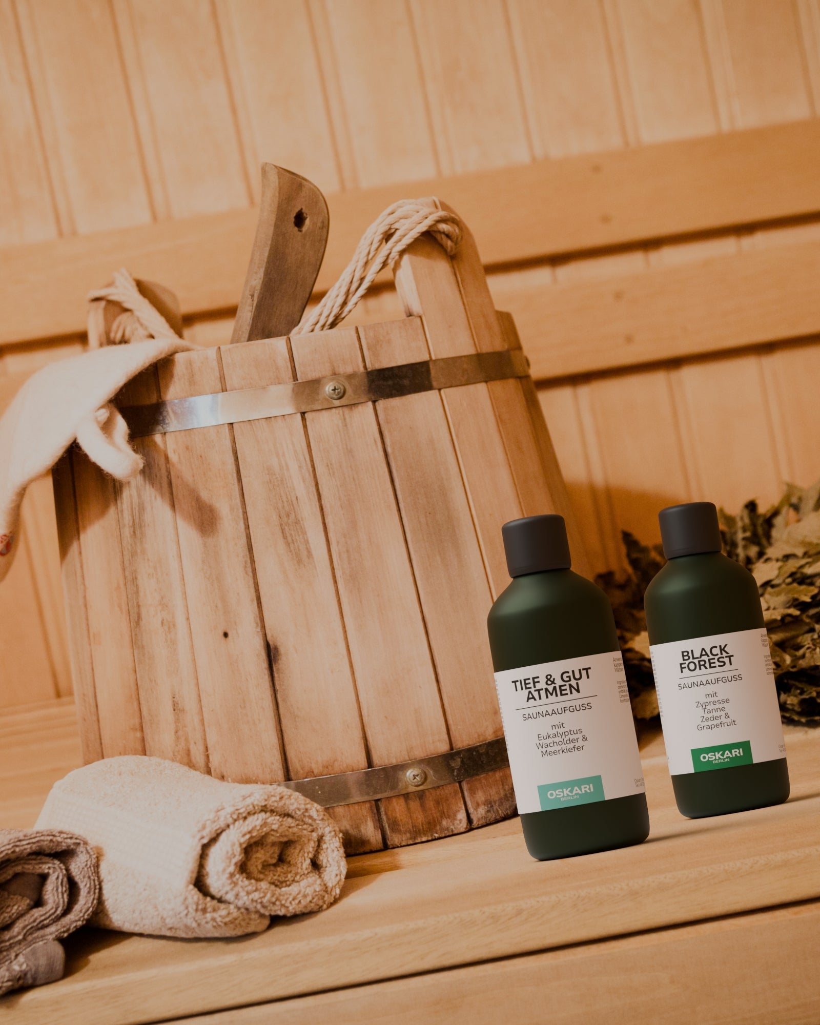 Zwei natürliche Saunaaufgüsse von Oskari stehen in einer auna aus Holz vor einem Saunaeimer und einem Saunahandtuch