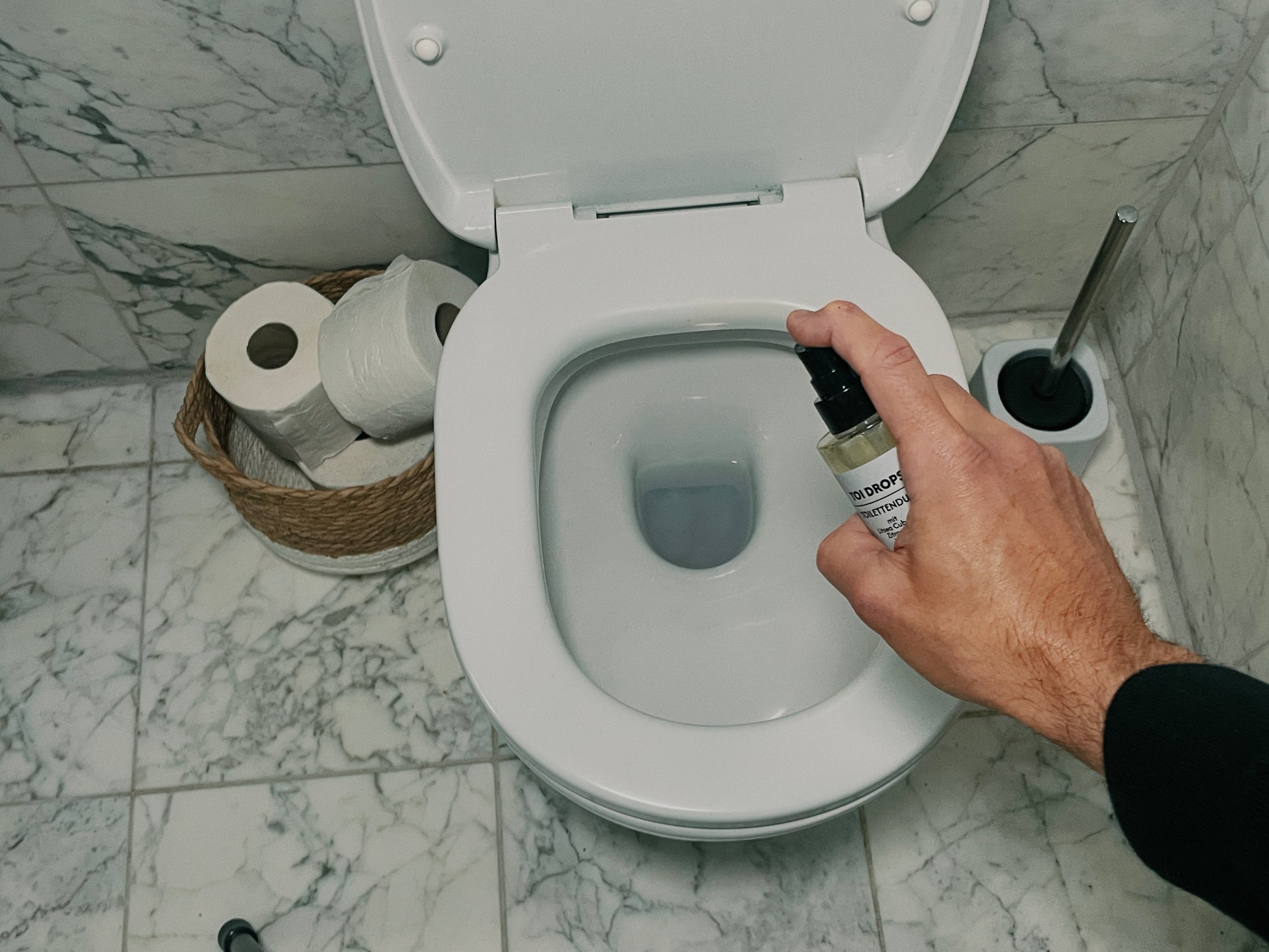 Eine Hand hält eine Toi Dros WC-Duft Flasche über eine geöffnete Toilettenschüssel