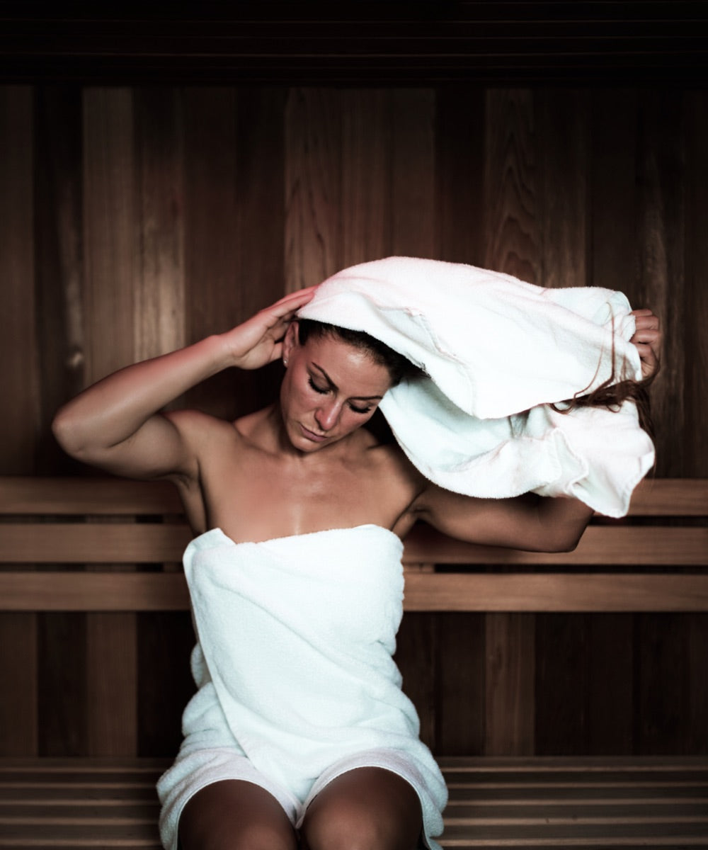 Eine Frau mit hellem Saunatuch um den Körper trocknet sich ihre Haare mit einem weißen Handtuch in einer Sauna