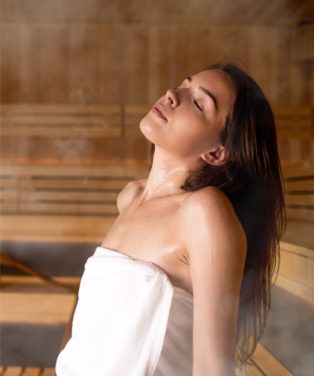 Ein junge Frau entspannt beim Saunieren und hat ein großes weißes Saunatuch um die Brust, Dampf steigt auf