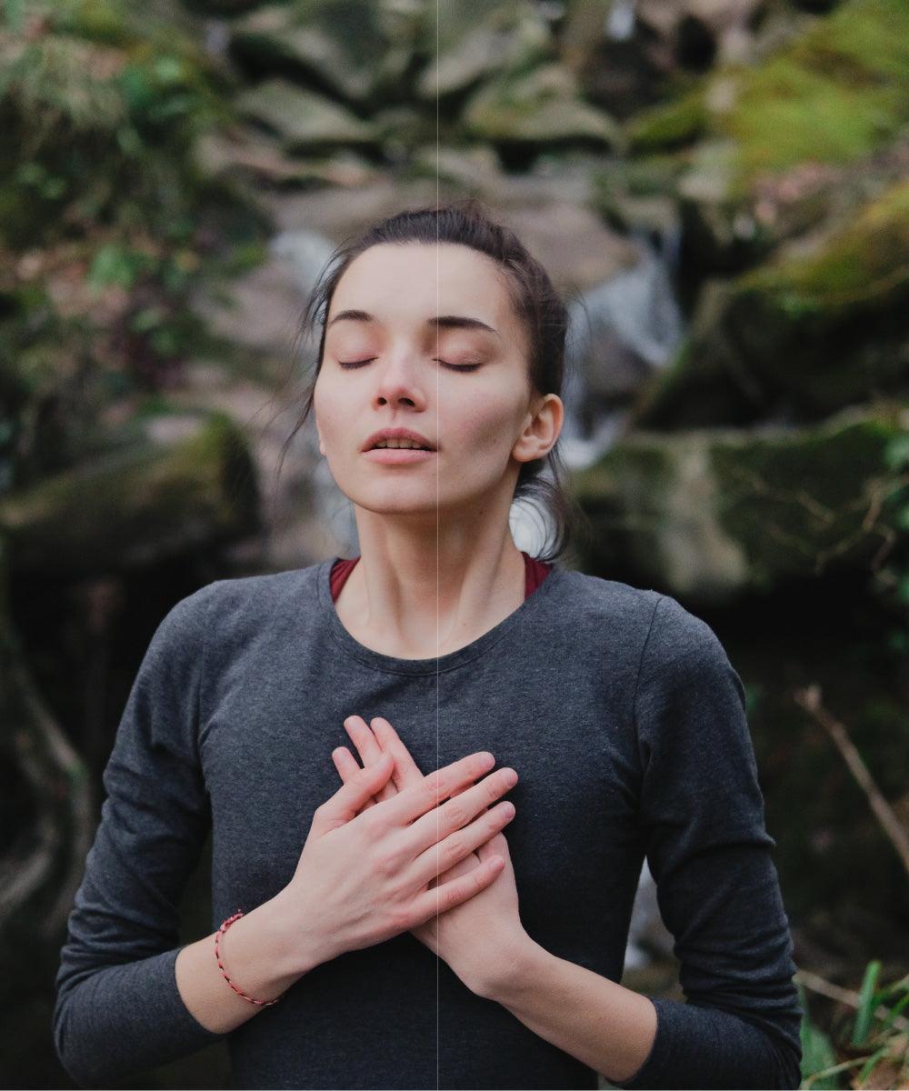 Junge Frau in grauem Pullover atmet tief durch in einer waldigen Umgebung mit Wasserfall im Hintergrund