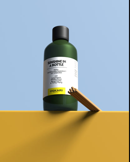 Auf einer warmgelben Anhöhe steht die Nachfüllflasche und die Duftstäbchen des Oskari Raumduftes mit Stäbchen Sunshine in a Bottle