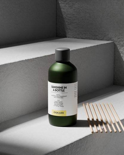 Die Refill-Flasche des Oskari Raumduftes Sunshine in a Bottle steht auf einer grauen Betontreppe, daneben liegend acht Papierstäbchen