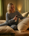 Junge Frau sitzt auf ihrem Bett in Nachtwäsche und benützt das Oskari Bio Kissenspray Bedtime Blend
