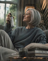Eine mittelate Frau mit kurzen grauen Haaren entspannt auf einem Sofa und sprüht das Bio Raumspray Chill Zone von Oskari