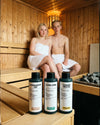 Junges Paar sitzt in einer Sauna hinter den drei Bio Saunaaufgüssen Alpen Zirbe, Entspannung Pur und Sommerfrische aus dem Bundle Trio von Oskari