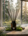 Der Raumduftflakon aus dunklem Glas von Oskari mit dem natürlichen Raumduft Breathe the Forest vor einem Fenster it Wald im Hintergrund